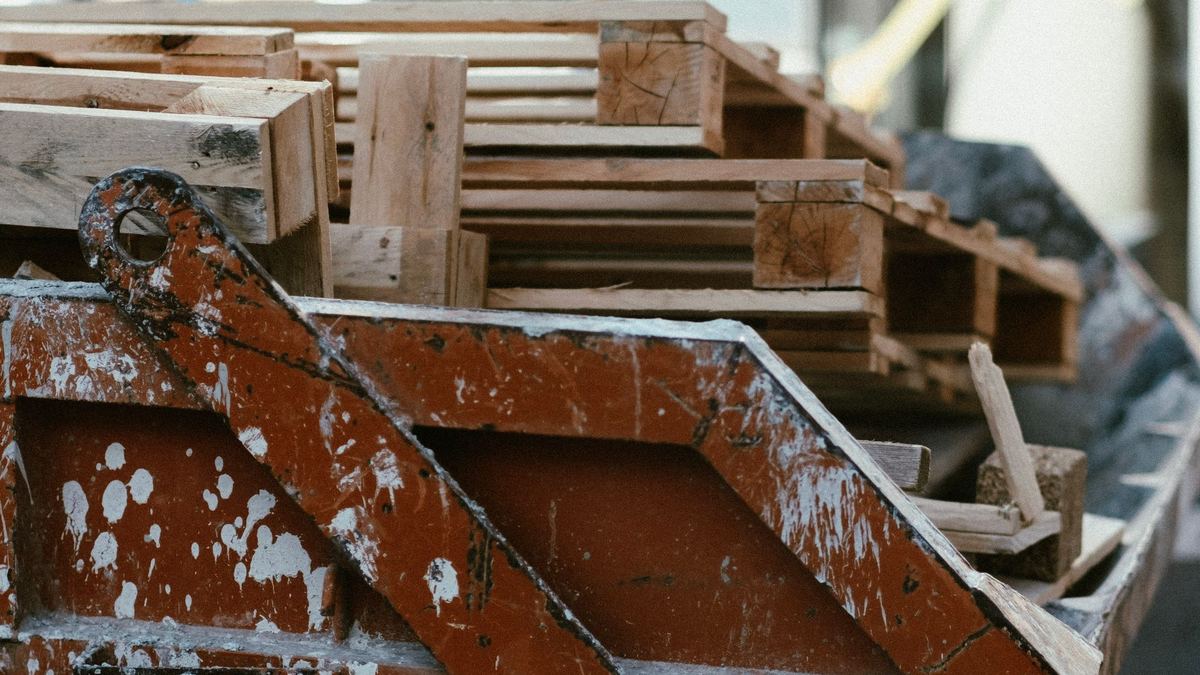 Caçamba metálica estacionária vista de perto, cheia de restos de madeira e pallets de obra, com a lataria pintada de vermelho desgastada